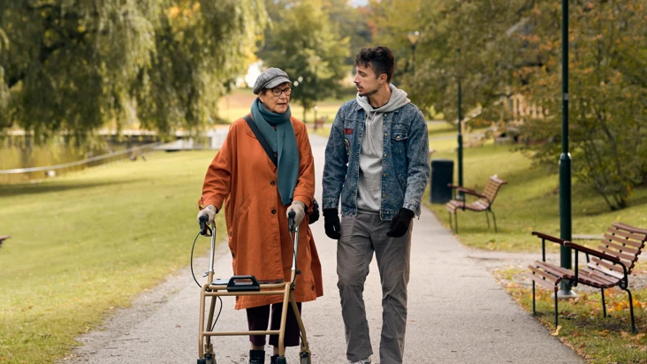 En &auml;ldre kvinna med rullator promenerar i en park tillsammans med en ung man.
